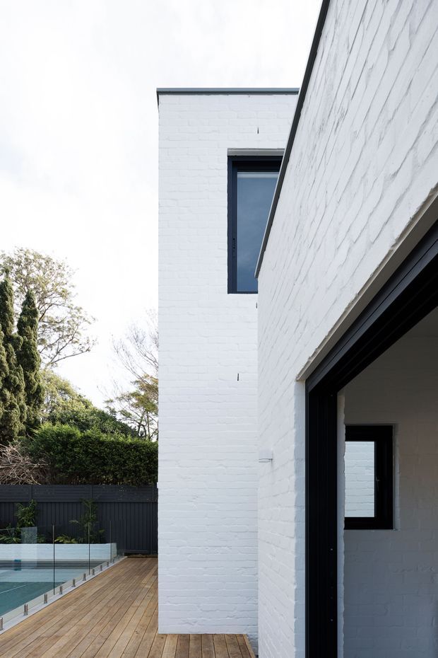 A narrow exterior view of a contemporary white-painted brick house, showing a tall two-storey wall with dark-framed windows and a large open doorway. A timber deck runs alongside the building, bordered by a frameless glass pool fence and dark privacy fencing, with landscaped vegetation beyond.
