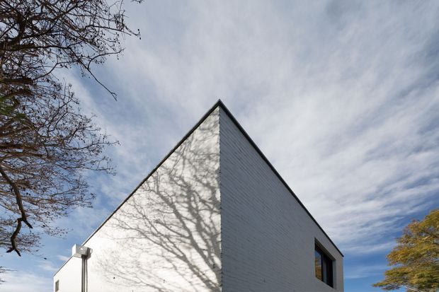 Exterior architectural photograph of a sharply pointed gable end on a simple white-painted masonry building. The triangular facade and adjoining side wall have a clean, minimal composition with a dark-framed rectangular window, while bare tree branches cast shadows across the facade. Blue sky and surrounding trees frame the structure.