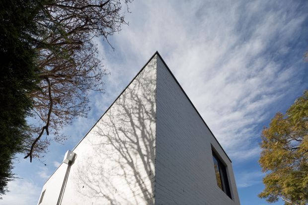 Low-angle exterior view of a sharply peaked, triangular-gabled building corner. The facade is clad in white horizontal weatherboard-like siding, with dark window and roofline trims. Bare tree branches cast shadows across the wall, with mature trees and blue sky surrounding the structure.
