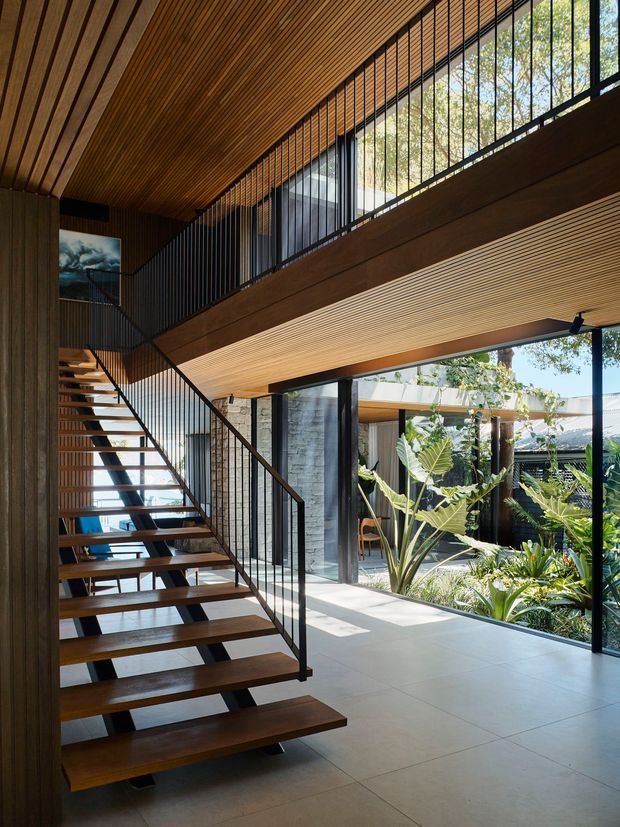 A double-height contemporary interior featuring an open-riser timber staircase with slender black metal balustrades, extensive floor-to-ceiling glazing, and a warm timber slatted ceiling. The stair and upper-level walkway overlook a bright open living area, with a planted courtyard visible through the glazing. The space combines strong indoor-outdoor connection, exposed structural beams, and abundant natural light.