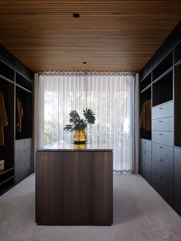 A sophisticated walk-in wardrobe with dark built-in cabinetry lining both sides, integrated drawers and hanging sections, and a central dark timber island with a reflective stone or glass top. A full-height sheer curtain filters daylight at the rear, while the ceiling is clad in narrow linear timber battens with recessed downlights. The space has a restrained, luxurious and warm contemporary appearance.
