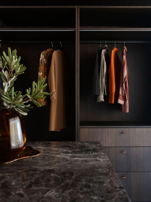 A dark, refined walk-in wardrobe with floor-to-ceiling built-in timber cabinetry, open hanging compartments, and drawers below. Several coats, jackets, and shirts hang inside the wardrobe. A dark veined stone island or bench occupies the foreground, topped with a sculptural plant in an amber glass vase. The mood is moody and luxurious, with warm timber tones and black interiors.
