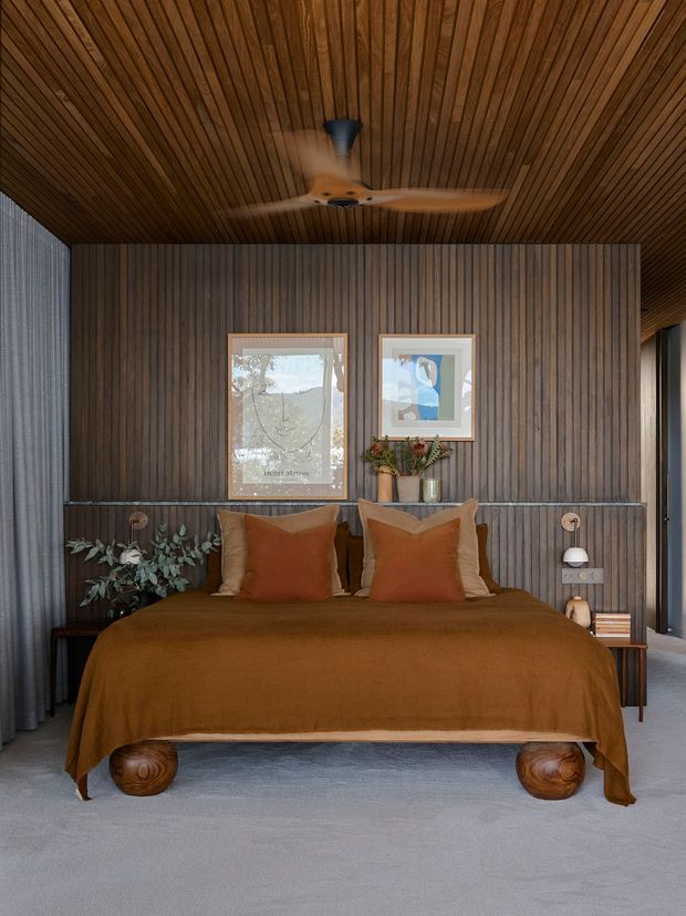 A warm, refined bedroom centered on a low platform bed with rust-brown bedding and layered cushions. The room features a dark timber slatted ceiling and matching vertical timber feature wall, grey carpet, full-height curtains, slim bedside tables, decorative wall-mounted reading lights, artwork, and sculptural timber bed supports. A ceiling fan is centrally mounted above the bed, and daylight enters through side glazing.