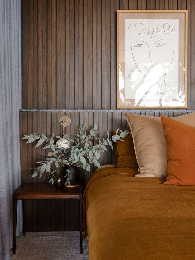 Close-up of a warmly styled bedroom corner featuring a bed with ochre-brown linen bedding and rust and tan cushions, set against a dark vertical timber-panelled wall. A small dark timber bedside table holds a vase of eucalyptus foliage and a simple lamp, while framed line-art adds a decorative focal point. Soft grey curtains and carpet provide neutral contrast.