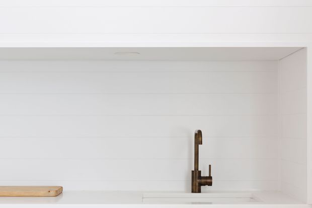 Minimalist white kitchen sink area with a white horizontal-panel splashback or wall finish, a pale timber cutting board at the left, and a dark bronze or aged-metal gooseneck tap centered over the undermount sink. The image is tightly framed with clean lines and bright, understated styling.