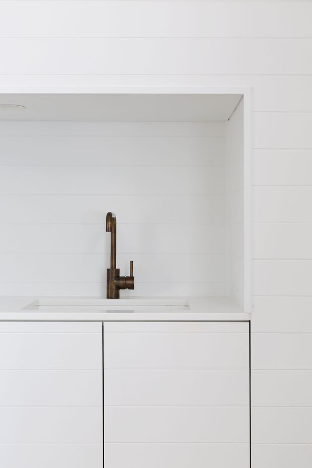 Minimalist close-up of a white built-in kitchen or utility sink set into streamlined white cabinetry. The recessed niche and surrounding walls are clad in horizontal white panelling, with a slim white benchtop and a distinctive brushed bronze or dark brass gooseneck mixer tap. The composition is highly pared back, bright, and monochromatic with warm metallic contrast.