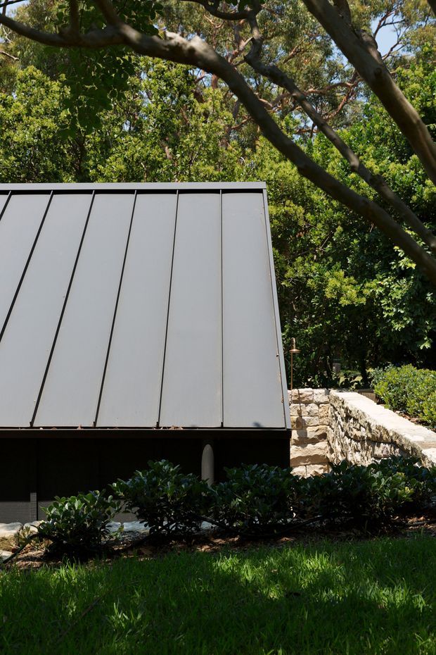 Exterior view of a compact modern structure set within dense landscaped greenery. The focal point is a steeply sloped standing-seam metal roof or wall plane in light grey, with dark lower cladding and a rough natural-stone retaining wall and pathway beside it. Mature tree branches frame the scene, with clipped shrubs and lawn in the foreground.