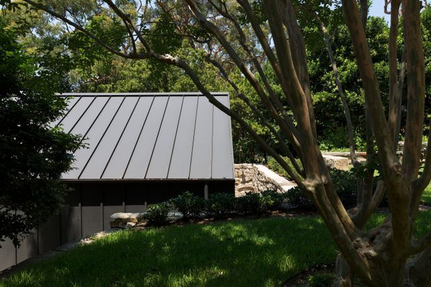 A low, modern garden structure is partially screened by mature trees and dense landscaping. The building has a simple dark lower volume topped by a prominent light grey standing-seam metal roof with strong linear panels and a steep single pitch. Sunlit lawn, rocks, shrubs and tree trunks surround the structure, creating a secluded landscaped setting.