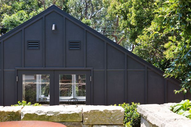 Front exterior view of a dark charcoal-clad building with a prominent steep gable roof and adjoining lower sloping roofline. Vertical panel detailing, small horizontal vents, an exterior wall light, and glazed double doors define the facade. A low rough-stone retaining wall and dense vegetation sit in the foreground and background.
