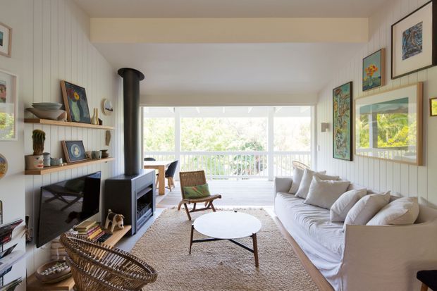 Bright, airy living room with a white slipcovered sofa, round coffee table, woven rug, black freestanding wood-burning stove, built-in open shelving, and extensive framed artwork. Large windows and glazed doors open toward a light-filled balcony or terrace, while vertical wall panelling and warm timber accents create a relaxed coastal character.