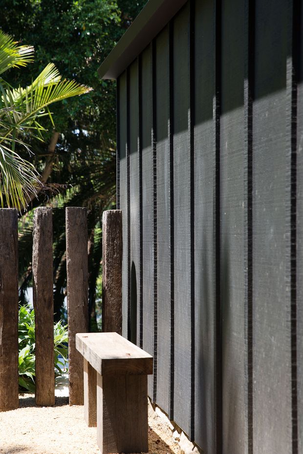 Close-up exterior view of a dark, vertically ribbed or battened facade beside a narrow gravel walkway. A simple raw timber bench and rough timber posts sit among lush tropical planting, with strong sunlight creating striped shadows across the wall. The image emphasises natural materials, texture, and the relationship between the building edge and garden.