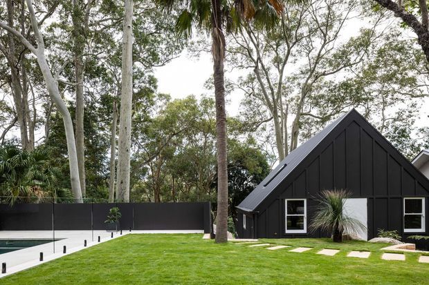 Exterior view of a dark, steeply gabled house or pavilion set within a landscaped garden. The black vertically clad facade has white-trimmed windows and a simple white door, with a manicured lawn, stepping stones, tall palms and established trees surrounding the building. A swimming pool and dark boundary wall are visible to the left.