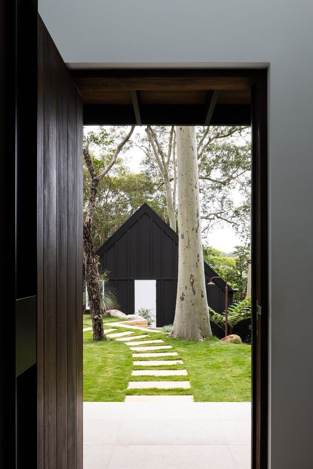 View from a dark timber-lined entryway through a large open doorway toward a landscaped garden. A stepping-stone path crosses the lawn to a small black, steep-gabled outbuilding, with mature trees and ferns surrounding the scene. The composition has a restrained, contemporary architectural character with strong contrast between the dark interior framing and bright green garden.