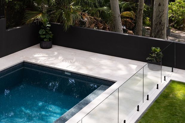 A compact modern landscaped pool area with a deep blue swimming pool edged in pale large-format paving. Frameless clear glass balustrades with dark metal fixings separate the pool terrace from a narrow lawn, while dark charcoal boundary walls and lush tropical planting create a private, contemporary outdoor setting.