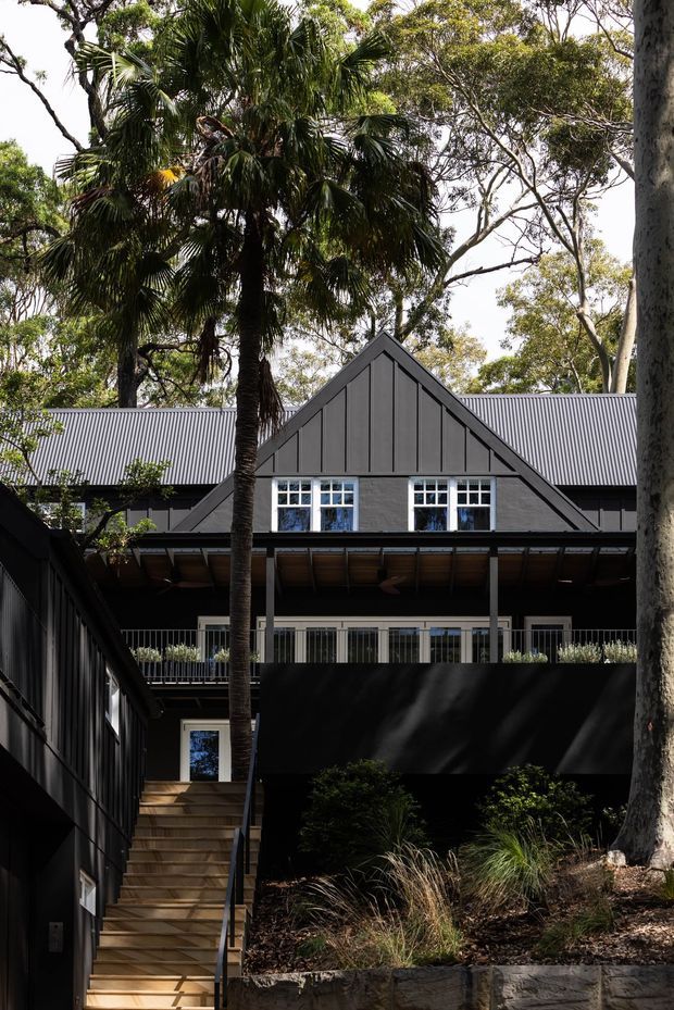 A multi-level dark-clad residential building set among mature trees and palms. The facade features a prominent central gable with vertical board cladding, a corrugated metal roof, white-framed multi-pane windows, covered balconies, and an external timber stairway. Native-style landscaped planting and stone retaining elements occupy the foreground.