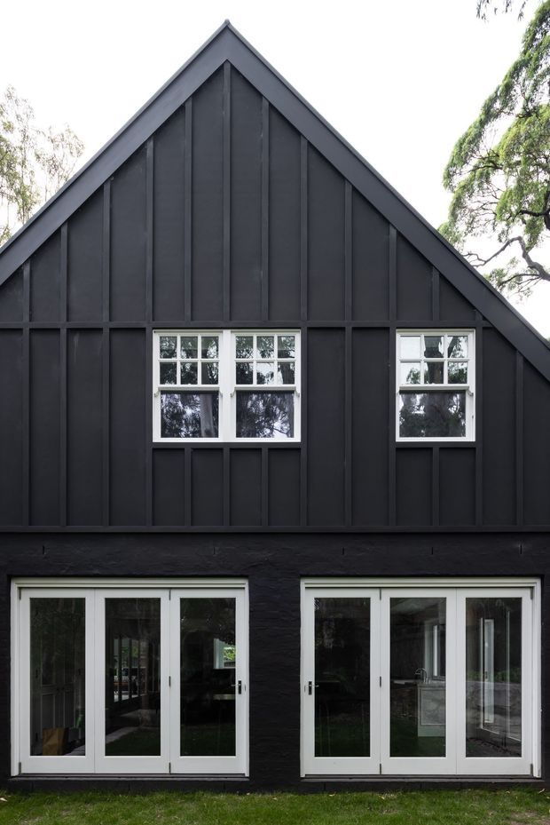 Front exterior view of a dark charcoal, steeply gabled building with a barn-like facade. The elevation features vertical board-and-batten cladding, white-trimmed multi-pane windows, and two sets of white-framed glazed double doors at ground level. Trees and lawn surround the building, while the glazing provides limited views into the interior.