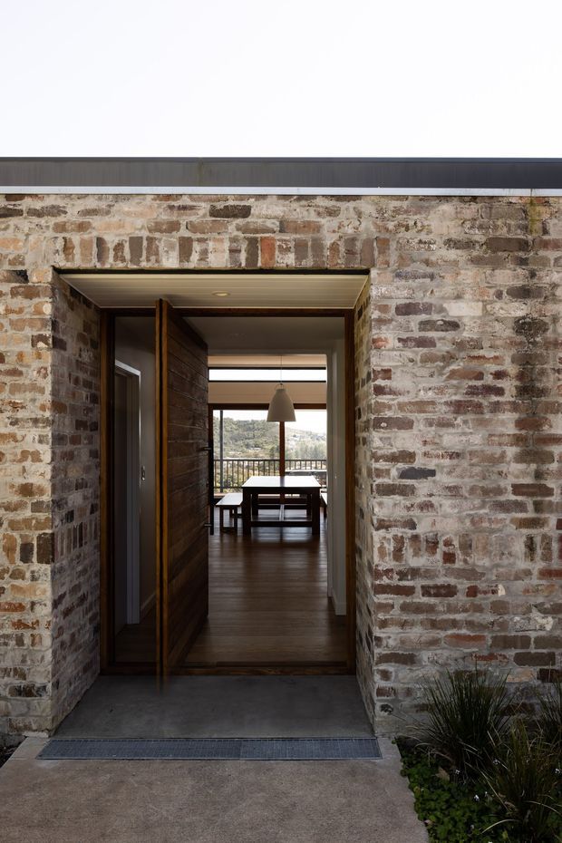 View toward the entrance of a contemporary brick-clad home. A recessed timber-lined doorway opens from a concrete threshold into a dark timber-floored interior, with a dining table and pendant light visible beyond. Full-height glazing at the rear frames a hilly landscape, while rough masonry and a restrained linear roof edge create a tactile, understated facade.