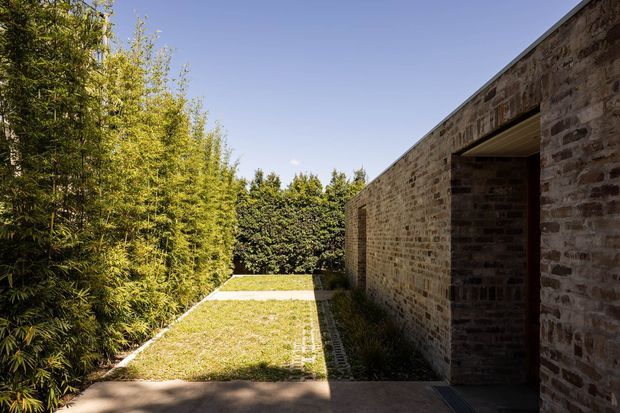 Exterior view of a low, rectilinear brick building opening onto a narrow landscaped courtyard or side garden. The facade is formed from warm weathered masonry with a deep-set doorway, while dense bamboo and clipped hedging create a private green edge. A simple paved path and lawn strip lead toward the rear, under clear daylight.