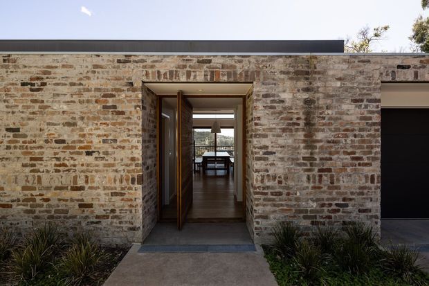 Front exterior view of a low, contemporary brick-clad residence with a flat roof and a deeply recessed timber-framed entry. The open doorway provides a sightline through the hall toward a dining or living area, while a dark garage door is visible at right. Native grasses and a concrete path frame the entry.