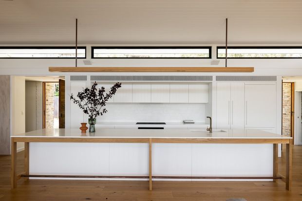Bright, refined contemporary kitchen viewed directly from the living area, featuring a large white island with a pale timber frame, integrated white cabinetry, a white stone-look benchtop, slim clerestory windows, timber flooring, and a long suspended timber light fitting. A vase with branches provides a natural decorative accent.