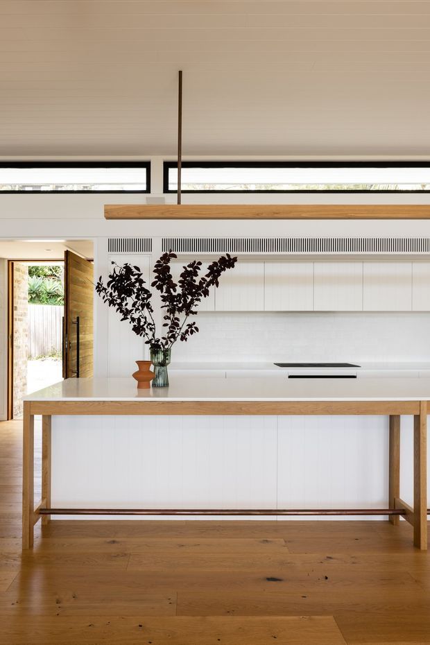 Bright contemporary kitchen with a long white island and integrated cooktop, pale vertical wall panelling, extensive white cabinetry, and narrow clerestory windows. A timber pendant light and timber island framing coordinate with the warm timber flooring. An open timber door leads outside, while a vase of dark leafy branches provides a restrained decorative accent.