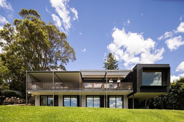 Exterior rear view of a contemporary two-storey home set in a landscaped suburban setting. The building has a long, rectilinear form with extensive glazing, a deep upper-level balcony with slender metal balustrading, pale rendered or concrete lower walls, and a contrasting dark-clad projecting volume at the right. A flat roof and broad roof overhangs reinforce the modern architectural character.