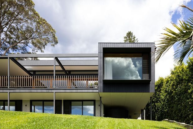 Exterior view of a contemporary multi-level residence on a sloping site, featuring a projecting dark-clad upper volume, extensive glazing, open balconies with slim metal railings, and a broad low roof or terrace structure. The home is surrounded by mature trees and dense landscaping under a partly cloudy sky.