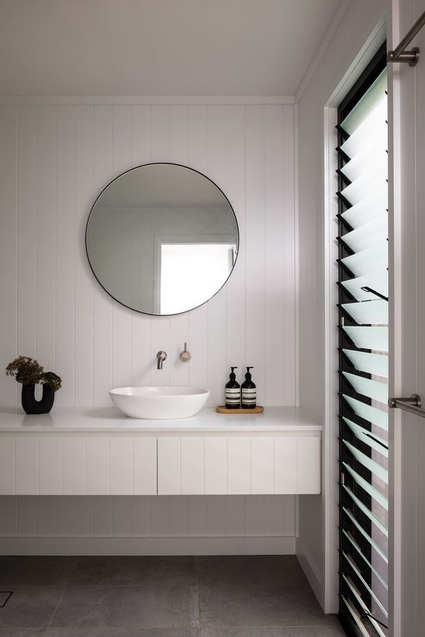 A bright, compact contemporary bathroom featuring a wall-mounted white vanity with an integrated vessel basin, round frameless mirror, white vertical wall panelling, dark large-format floor tiles, and a tall louvred window or door providing filtered daylight. Minimal black and neutral accessories sit on the vanity.