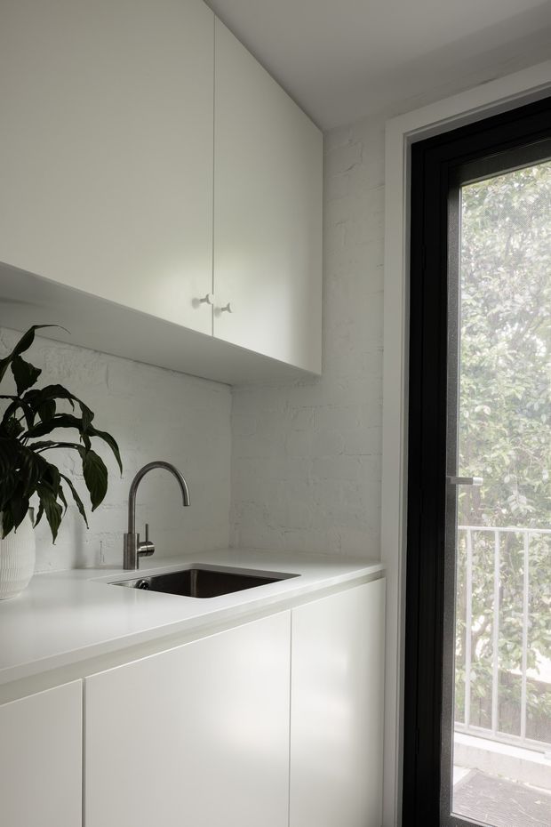 A compact, minimalist white kitchen or kitchenette with seamless flat-panel cabinetry, a white countertop, undermount sink and curved chrome tap. The backsplash and surrounding walls are painted white brick, while a black-framed glazed door provides daylight and views of greenery. A potted plant adds a small natural accent.