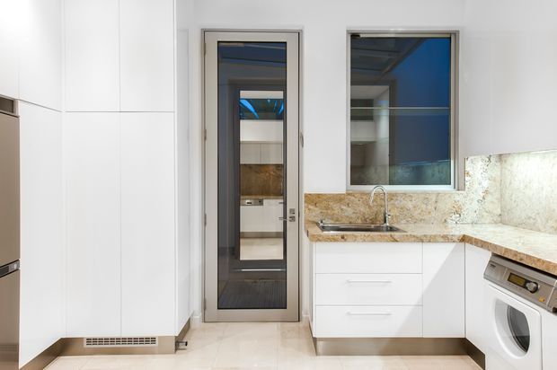 Bright contemporary kitchen and laundry area with white full-height cabinetry, pale tiled flooring, a warm mottled stone benchtop and splashback, stainless-steel sink and dishwasher, and a front-loading washing machine. A glazed internal door and window bring in views and reflected blue tones.