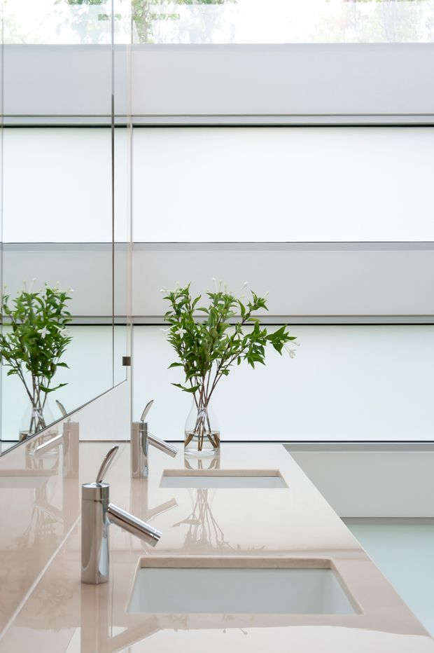 A bright, minimalist bathroom vanity with a long pale stone or engineered-stone benchtop, two rectangular integrated basins, and sleek chrome single-lever faucets. A large frameless mirror reflects the vanity and small glass vases of leafy branches. The background features translucent horizontal glazing or wall panels, creating a softly illuminated, contemporary interior.