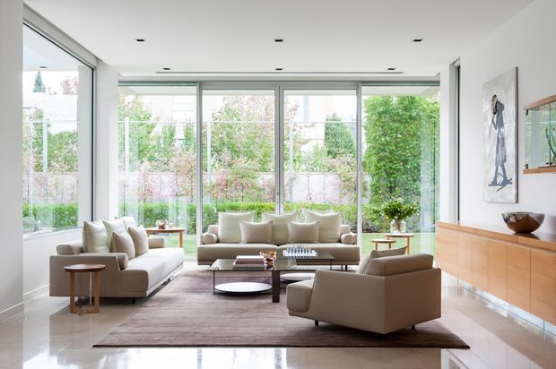 Bright, refined living room with floor-to-ceiling glazing on multiple sides, a neutral sofa and armchairs arranged around layered coffee tables, a large area rug, and a long timber-toned built-in sideboard. The space has a clean, contemporary architectural shell with recessed ceiling lighting and views to landscaped greenery outside.