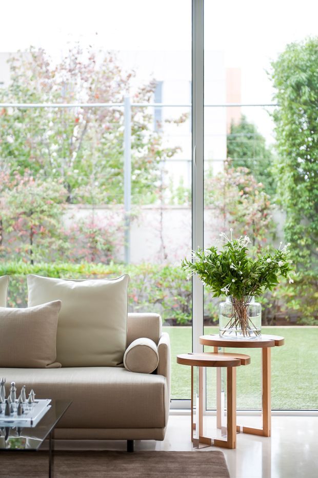 Bright, refined living room corner with a beige upholstered sofa, layered neutral cushions, nested wood-and-glass side tables, and a glass vase of greenery. Floor-to-ceiling glazing opens the room visually to a landscaped garden, while a dark glass coffee table and soft area rug add contrast. The space has a calm, contemporary feel with abundant natural light.