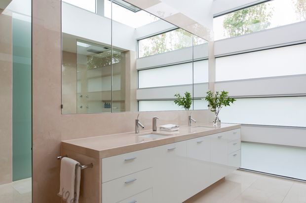 Bright contemporary bathroom with a large floating white vanity, pale stone benchtop, twin basins and chrome tapware. Full-height mirrors reflect a glass shower enclosure, while extensive clerestory glazing and translucent horizontal panels provide abundant natural light. The room has a spacious, clean and refined appearance.