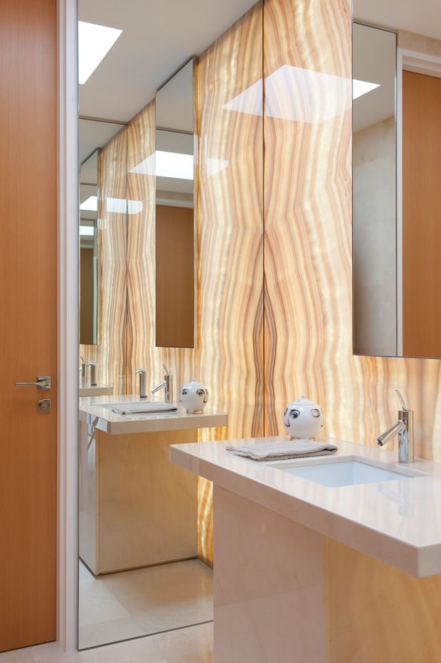 Contemporary bathroom vanity with a floating white benchtop, integrated basin, chrome tapware, light timber cabinetry, full-height mirrors, and a dramatic illuminated vertically veined stone feature wall. A timber door and reflected vanity extend the compact, refined space.