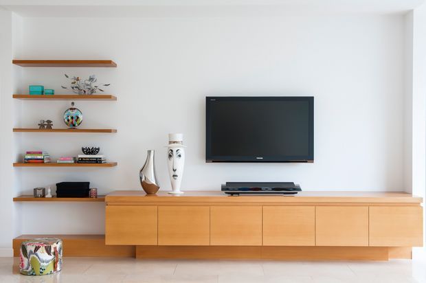 Bright minimalist living room with a long wall-mounted timber media console, flat-screen television, open built-in display shelving, and carefully arranged decorative objects. The white walls and pale floor create a clean, contemporary interior.