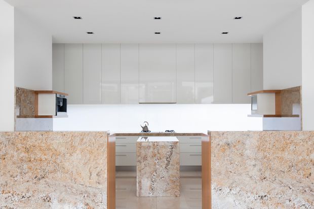 Bright, minimalist contemporary kitchen viewed straight on, featuring a central island, extensive glossy white full-height cabinetry, white splashback, integrated appliances, and prominent warm beige-and-brown natural stone benchtops and side panels. Recessed ceiling lights and pale flooring create a clean, refined appearance.