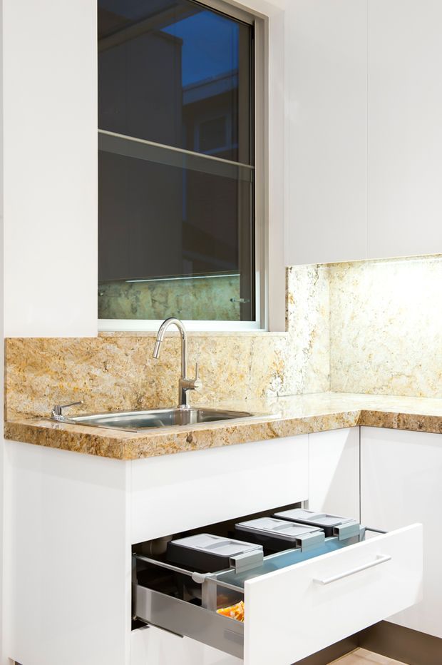 A bright, compact contemporary kitchen corner featuring white cabinetry, a stainless-steel sink with a tall curved faucet, and warm brown-gold natural stone benchtops and splashback. A large dark-framed window sits above the sink, while a deep lower drawer is open to reveal organised storage bins.
