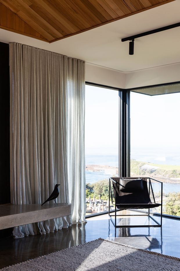 A refined coastal living space with expansive corner glazing overlooking a rocky shoreline and water. The room features a warm timber-lined ceiling, pale plaster walls, dark-framed windows, full-height pleated neutral curtains, polished dark flooring, a minimalist black sling chair, a low concrete or stone console, and a textured neutral rug. A black track light and sculptural bird ornament add restrained contemporary detail.