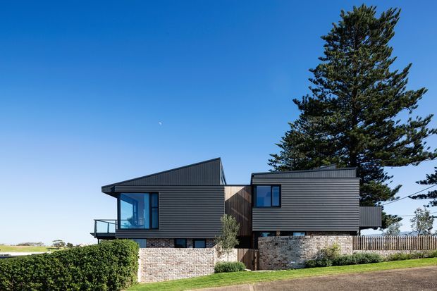 Exterior view of a contemporary multi-level residential house with two offset dark horizontal-clad volumes, contrasting timber-toned central infill, broad glazing, and a low brick retaining wall. The home sits in a landscaped suburban or coastal setting with mature evergreen trees and a clear blue sky.