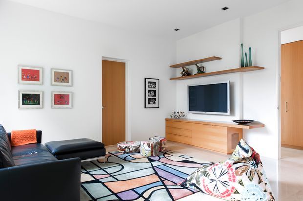 Bright contemporary living room with white walls, a black leather sectional and ottoman, colourful patterned cushions, and a large geometric multicoloured area rug. A timber media console with wall-mounted television and floating timber shelves forms the main feature wall, with framed artwork and timber interior doors adding warmth.