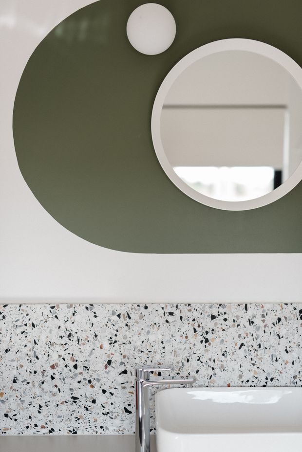 Close-up view of a contemporary bathroom vanity area featuring a large round mirror, a smaller round wall light, an olive-green organic-shaped painted wall panel, terrazzo wall cladding, and a chrome tap beside a white basin.