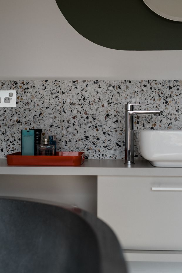 Close-up view of a contemporary bathroom vanity with a white vessel basin, chrome mixer tap, pale cabinetry, and a terrazzo-style speckled backsplash. Toiletries sit in a red tray on the benchtop, with a partial round mirror visible above.