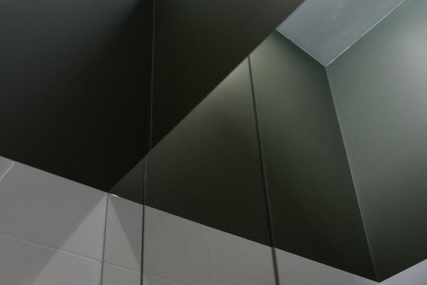 Abstract close-up of a dark, angular architectural ceiling or overhead surface meeting pale tiled walls. Sharp geometric planes, narrow dark seams, and contrasting charcoal and light grey finishes create a highly minimalist, sculptural composition.