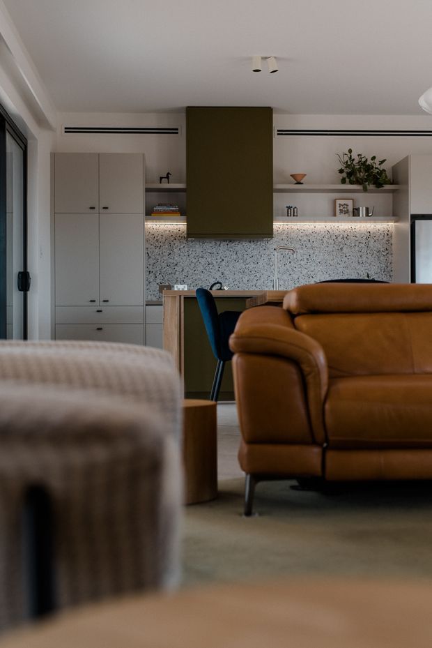 Interior view of an open-plan living and kitchen area. A warm cognac leather sofa dominates the foreground, with a softly upholstered chair and dining seating nearby. The kitchen features pale built-in cabinetry, a dark central rangehood, open shelving, a terrazzo-style speckled splashback, timber island cabinetry and under-shelf lighting. The space has a restrained, warm contemporary feel with concealed linear air vents and understated ceiling spotlights.