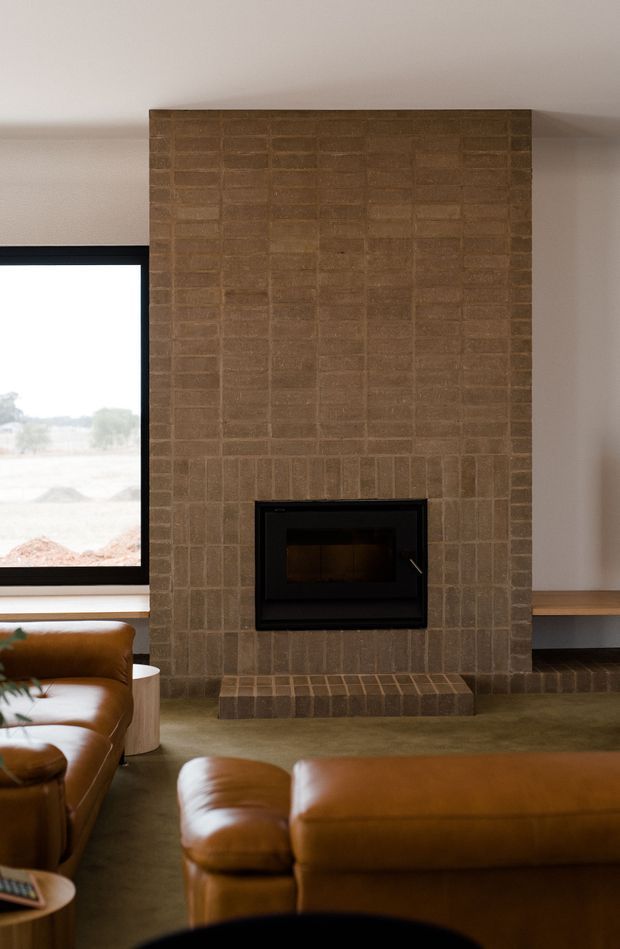 A warm, minimalist living room centred on a tall textured brown brick fireplace with a black inset wood-burning firebox. A large black-framed window sits beside the fireplace, while caramel-brown leather sofas, a muted green carpet and simple timber shelving create a refined earthy interior.