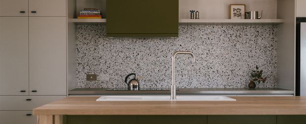 A compact contemporary kitchen viewed straight on, featuring a long timber island with an undermount sink and tall brushed-metal tap, dark green lower cabinetry, pale full-height cabinetry at left, and an open shelf above the benchtop. The backsplash is a distinctive terrazzo-style surface with dense dark and light aggregate, while restrained shelving decor and warm muted lighting create a refined, softly contrasting interior.