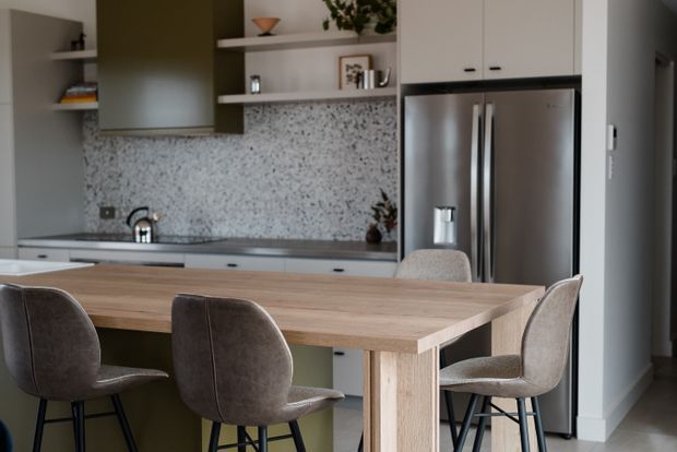 A contemporary open-plan kitchen and dining area with a light timber dining table and upholstered bar stools in the foreground. The kitchen features pale cabinetry, integrated open shelving, a stainless-steel double-door refrigerator, dark benchtops, and a speckled terrazzo-style splashback. Neutral grey, white, and timber tones create a refined, understated interior.
