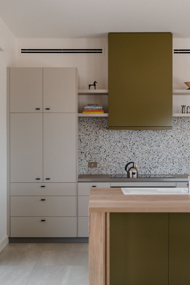 A compact contemporary kitchen with full-height pale grey cabinetry, an olive-green rangehood enclosure and matching island joinery, a terrazzo-style speckled backsplash, integrated stainless-steel sink and tap, timber island benchtop, and large light grey floor tiles. Open shelving displays a few books and objects, while linear black ventilation grilles add a graphic detail.