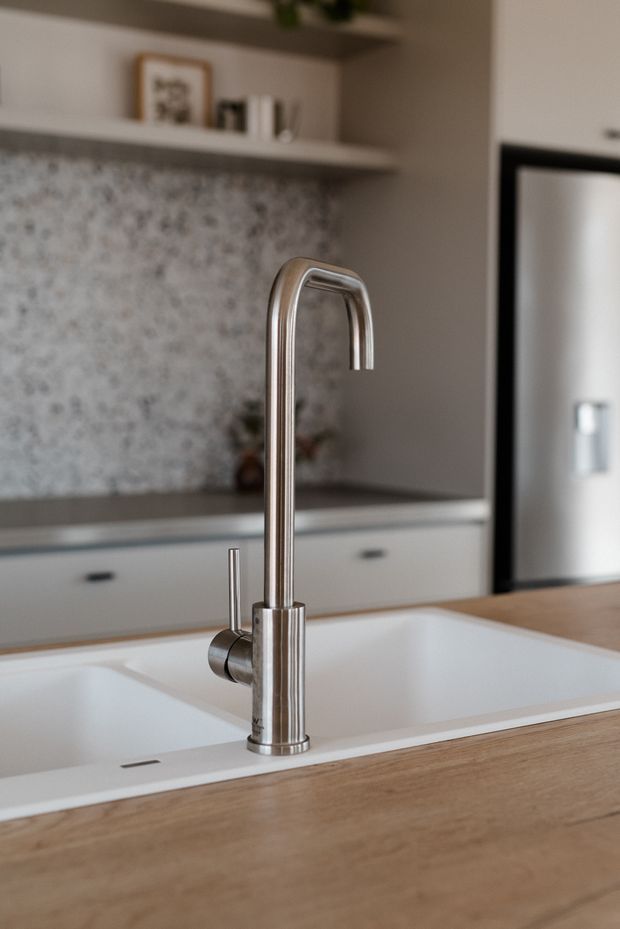 Close-up view of a contemporary kitchen sink and tall curved gooseneck mixer tap set into a timber-toned island benchtop. The kitchen features white cabinetry with dark handles, a mottled stone or terrazzo-style splashback, open shelving, and a partially visible black-framed doorway or window. The image uses shallow depth of field and warm, soft natural lighting.